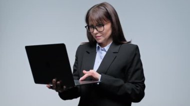 Orta yaşlı ciddi bir iş kadını el ele tutuşmuş, gri bir geçmişi var. İş için dizüstü bilgisayar kullanan, gözlüklü, siyah takım elbiseli olgun bir kadın internet teknolojisi sunuyor.