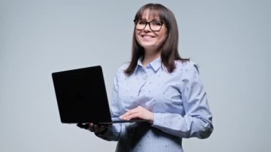 Orta yaşlı gülümseyen iş kadını gri arka planda dizüstü bilgisayarını tutuyor. Mavi gömlekli neşeli olgun kadın kameraya bakıyor. Çevrimiçi hizmet, iş işi, internet teknolojisi