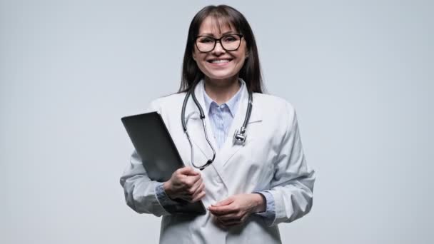 灰色の背景にラップトップコンピュータを保持する中年の笑顔の女性医師 カメラを見てメガネで白いコートの正の医師 ヘルスケア オンライン医療サービス — ストック動画