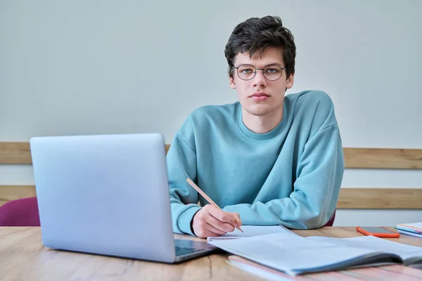 年轻的大学生坐在课桌前 用笔记本电脑 笔记本写作 看着相机 语言课程 青年概念 — 图库照片