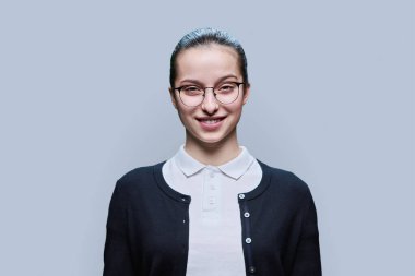Gri stüdyo geçmişinde mutlu lise öğrencisinin yakın plan portresi. Dişleriyle gülümseyen, gözlüklü genç bir kadın kameraya bakıyor. Ergenlik, yaşam tarzı eğitimi boş zamanlardaki gençler.