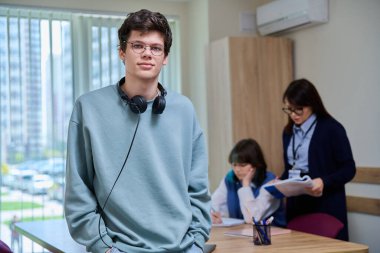 Sınıfın içinde kameraya bakan üniversite öğrencisinin portresi. 18-19 yaşlarında, gözlüklü, kulaklıklı, eğitim merkezinde poz veren genç bir erkek. Eğitim, bilgi, öğrenme, gençlik