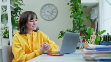 Neşeli genç kız kolej öğrencisi masasında oturmuş, gülüyor bilgisayarın kamerasına bakıyor ve sohbet konferansı yapıyor. Boş zaman çalışması için çevrimiçi internet teknolojisi