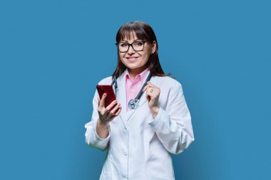 Mavi stüdyo arka planında telefon kullanan beyaz önlüklü olgun bir kadın doktor. Tıp, internet hizmeti, meslek, sağlık, sağlık hizmetleri, profesyonel mobil uygulamalar, tedavi kavramı