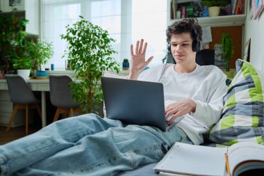 Kulaklıklı genç adam bilgisayarı görüntülü konuşma, çalışma, boş zaman, evdeki kanepede uzanmak için kullanıyor. İnternet teknolojisi, sanal toplantılar, modern yaşam tarzı, e-öğrenme, gençlik koleji yaşı
