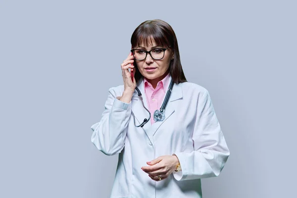 身穿白色实验室外套的成熟女医生在电话交谈 背景是灰色的工作室 治疗概念 — 图库照片