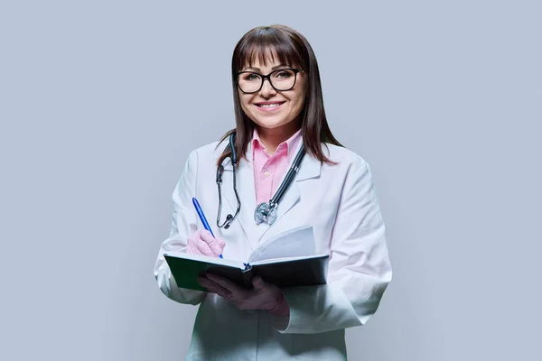 穿着白色实验室外套的成熟女医生微笑 注意工作笔记本上的信息 看着灰色工作室背景的相机 医疗服务职业卫生保健治疗 — 图库照片