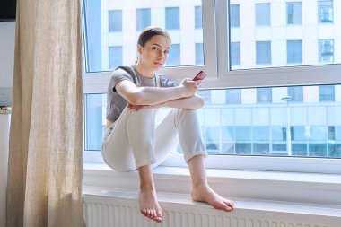 Pencere kenarında oturan genç kız elinde akıllı telefonuyla kameraya bakıyor. Sosyal ağlarda sohbet eden genç bir kadın. İzleme, mobil uygulamalar, internet teknolojileri.