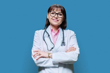 Mavi arka planda çapraz kollu beyaz önlüklü, kendine güvenen olgun kadın doktor portresi. Gülümseyen orta yaşlı kadın kameraya bakıyor. Meslek hekimliği mesleği sağlık hizmetleri