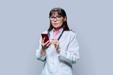 Beyaz laboratuvar önlüklü olgun bir bayan doktor, gri stüdyo geçmişinde telefon kullanıyor. Tıp, internet hizmeti, meslek, sağlık, sağlık hizmetleri, profesyonel mobil uygulamalar, tedavi kavramı