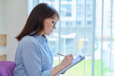 Orta yaşlı, ciddi bir kadın. Pencere kenarındaki ofiste panosu var. Kağıt belgeli iş kadını, el yazısı notlar, profil görünümü, kopyalama alanı