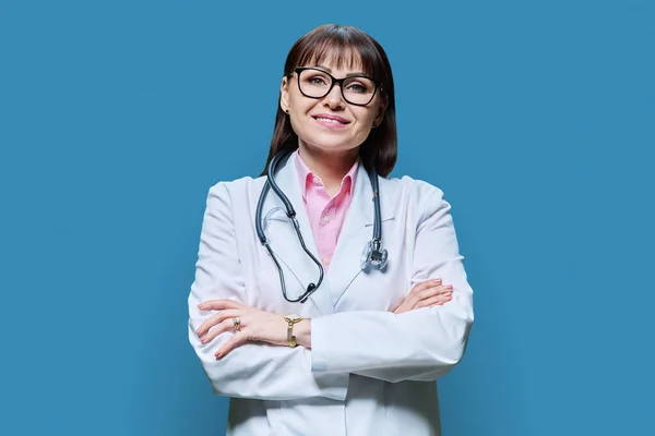 有自信的成熟女医生 身穿白衣 双臂交叉 背景为蓝色 在镜头前微笑的中年女性 职业医学职业健康保护卫生保健 — 图库照片