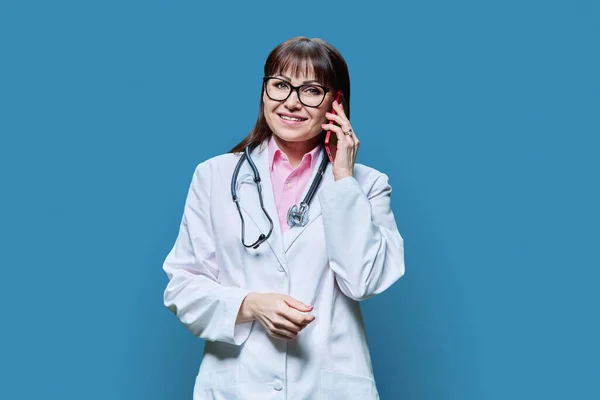 身穿白色实验室外套的成熟女医生在电话交谈 背景为蓝色 治疗概念 — 图库照片