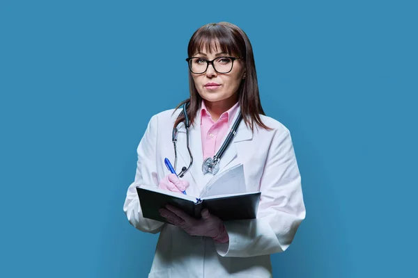 严肃而成熟的女医生穿着白色的实验室外套 记录工作笔记本上的信息 看着蓝色工作室背景的相机 医疗服务职业卫生保健治疗 — 图库照片