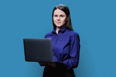 Mavi stüdyo arka planında dizüstü bilgisayarla poz veren genç iş kadını. Siyah tişörtlü, gülümseyen bir kadın kameraya, iş eğitimine, dijital internet teknolojisine, insanlara bakıyor.
