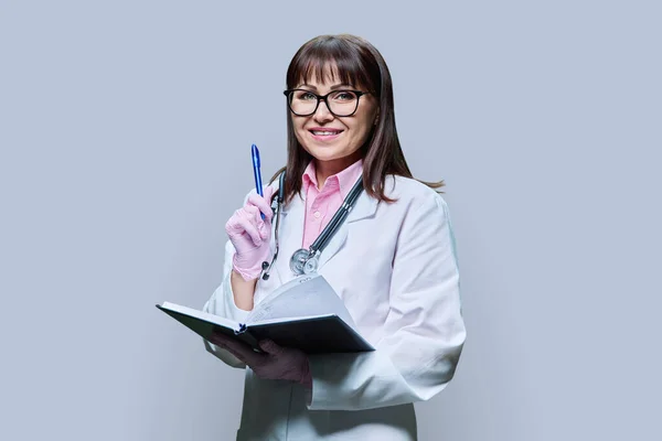 穿着白色实验室外套的成熟女医生微笑 注意工作笔记本上的信息 看着灰色工作室背景的相机 医疗服务职业卫生保健治疗 — 图库照片