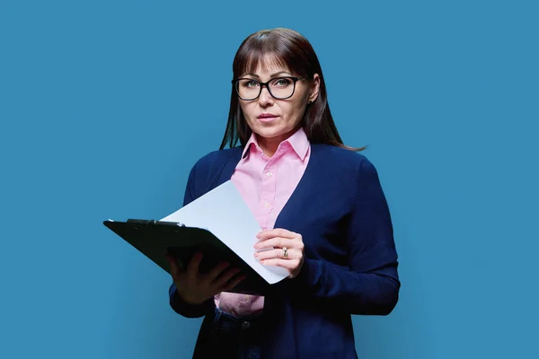 Serious mature business woman with clipboard looking at camera on blue background. Business, work, job, control, audit, statistics, inspection concept