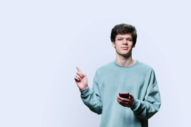 Elinde akıllı telefon olan genç bir üniversite öğrencisi mesaj, beyaz arka plan ve fotokopi için parmağıyla yeri gösteriyor. Gençlik eğitimi eğlence kavramı için reklam, tavsiye, mal hizmetleri
