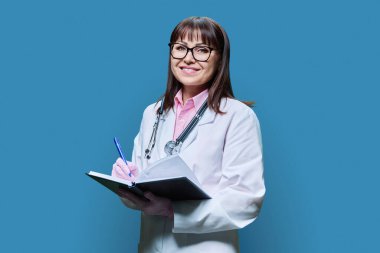 Beyaz laboratuvar önlüklü, gülümseyen olgun bayan doktor çalışan not defterine not alıyor, mavi stüdyo arkaplanındaki kameraya bakıyor. İlaç, hizmet, meslek, sağlık, sağlık hizmetleri