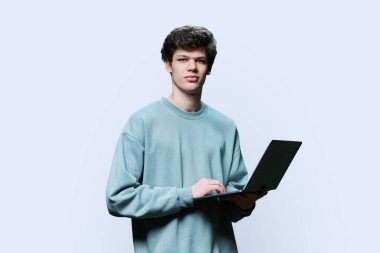 Laptop kullanan genç bir üniversite öğrencisi, beyaz stüdyo geçmişi var. Kameraya bakan gülümseyen yakışıklı adam. İnternet teknolojileri, çevrimiçi öğrenim istirahatı, sanal uzaktan eğitim eğlence iletişimi