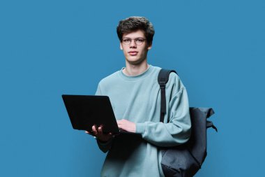 Laptop kullanan genç bir üniversite öğrencisi, mavi stüdyo geçmişi var. Kameraya bakan gülümseyen yakışıklı adam. İnternet teknolojileri, çevrimiçi öğrenim istirahatı, sanal uzaktan eğitim eğlence iletişimi