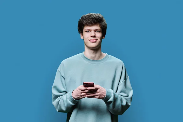 Młody Uśmiechnięty Student Koledżu Smartfonem Patrzący Kamerę Niebieskim Tle Przystojny — Zdjęcie stockowe