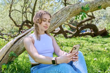 Parktaki çimlerin üzerinde oturan genç bir kadın akıllı telefonuyla rahatlıyor. Boş zaman, öğrenim, alışveriş, rahatlama için mobil uygulamalar, çevrimiçi internet teknolojisi