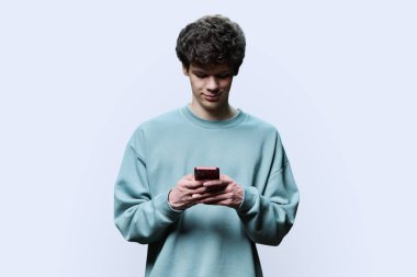 Beyaz arka planda akıllı telefona bakan gülümseyen genç bir üniversite öğrencisi. Kıvırcık saçlı, yakışıklı genç bir erkek. İnternet teknolojileri, eğlence öğrenme hizmetleri için mobil çevrimiçi uygulamalar