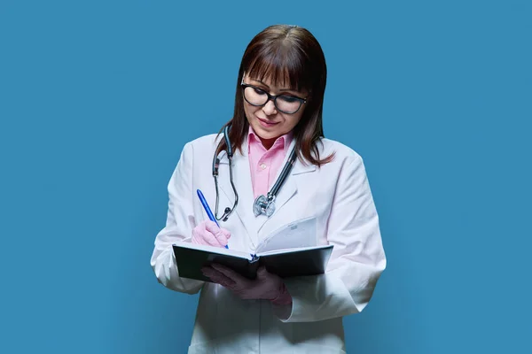 穿着白色实验室外套的成熟女医生 记录工作笔记本上的信息 有蓝色工作室背景 服务业 医疗保健 医疗保健 — 图库照片