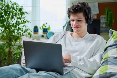 Kulaklıklı genç adam evdeki kanepede dizüstü bilgisayar kullanarak yatıyor. Gülümseyen erkek çalışma, dinlenme, iletişim, internet teknolojisi, modern yaşam tarzı