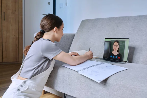 stock image Teenage girl studying math looking at laptop with online lesson, teacher on screen, making notes in notebook, from home. Internet online video technology in teaching education, virtual remote classes