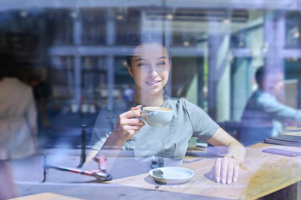 窓ガラス越しにカメラを眺めながらコーヒーを飲みながらカフェの中で美しい若い女性 カフェで休んでいる魅力的な10代の女の子 ライフスタイルの概念 — ストック写真