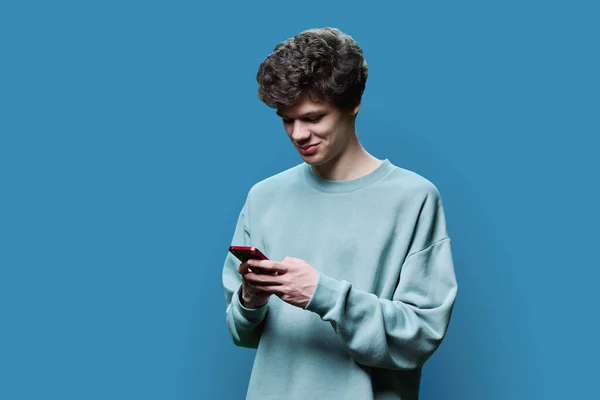 Młody Uśmiechnięty Student Patrzący Smartfona Niebieskim Tle Przystojny Nastolatek Kręconą — Zdjęcie stockowe