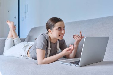Evdeki genç kız kanepede uzanıyor, duygusal konuşmalar dizüstü bilgisayardaki webcam 'e bakıyor, görüntülü sohbet görüşmesi yapıyor. Boş zaman iletişimi, modern yaşam tarzı, ergenlik kavramı öğrenme teknolojisi