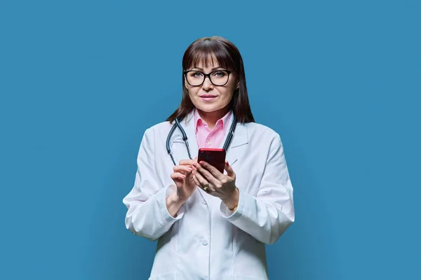 Médica Madura Jaleco Branco Usando Telefone Fundo Estúdio Azul Medicina — Fotografia de Stock