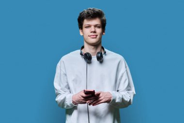 Mavi stüdyo arka planında akıllı telefon tutan kulaklıklı yakışıklı bir erkek. 18-19 yaşlarında üniversite öğrencisi, kameraya bakıyor. İnternet teknolojisi, mobil uygulamalar