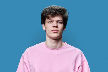 Mavi stüdyo arka planındaki kameraya bakan genç adamın portresi. Kendine güvenen 18-19 yaşlarında, pembe tişörtlü kıvırcık saçlı bir erkek. Üniversite öğrencisi, yaşam tarzı, eğitim konsepti