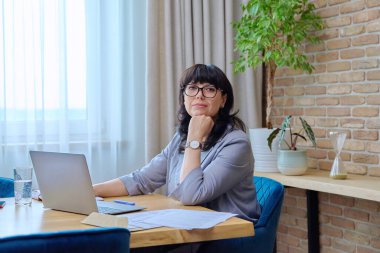 Kendine güvenen, gülümseyen, olgun bir kadın, dizüstü bilgisayarla iş sözleşmesi yapıyor, ofisteki kameralara bakıyor. 50 'lerin kadın lider yöneticisi finans müdürü iş yeri