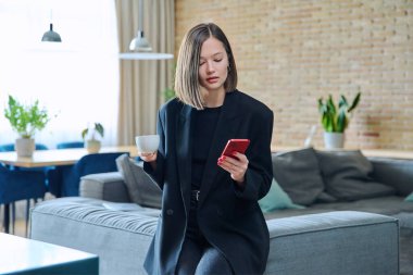 Genç, modaya uygun, güzel bir kadın evinde, elinde bir fincan kahveyle akıllı telefon kullanıyor. İnternet teknolojisi, gezici uygulamalar, boş zaman işi alışverişi.