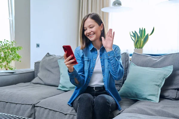 비디오 콘퍼런스를 여성은 집에서 소파에 앉아서 스마트폰으로 흔들며 인사를 생활을 — 스톡 사진