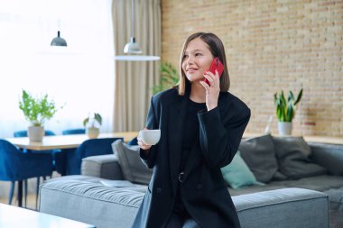 Genç, modaya uygun, güzel bir kadın oturma odasında kanepede oturuyor cep telefonuyla konuşuyor, rahatlıyor, bir fincan kahveyle. Yaşam tarzı, eğlence, ev dekorasyonu, gençlik konsepti.