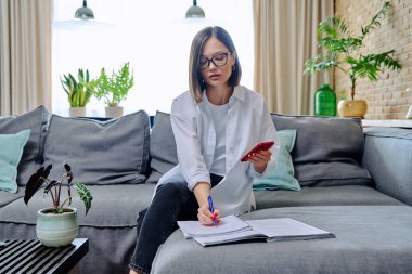 Genç kız üniversite öğrencisi evde oturuyor, kanepeye oturuyor, oturma odasında, defterde akıllı telefon yazıları kullanıyor. Eğitim bilgisi gençlik mobil uygulamaları İnternet çevrimiçi öğrenim hizmetleri