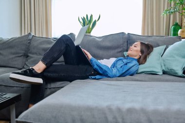 Genç bir kadın dizüstü bilgisayar kullanarak kanepede yatıyor. Çalışmak için internet teknolojileri, uzaktan çalışan kadın serbest çalışan, online ders izleyen üniversite öğrencisi, sosyal ağlarda dinlenme iletişimi