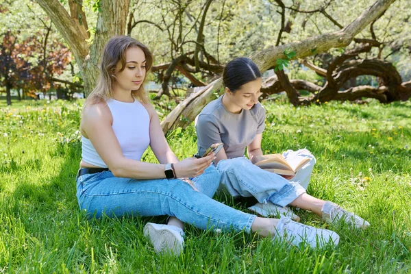 Две Подружки Подростки Отдыхают Траве Используя Книгу Чтения Смартфонов Весна — стоковое фото