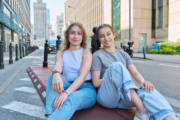 stock image Two smiling teenage female students looking at camera, modern urban style, buildings background. Fashionable girls sitting on sidewalk near road. Youth, fashion, style, city, lifestyle concept