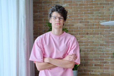 18-19 yaşlarında ev içinde çapraz kollu yakışıklı bir adamın portresi. Gözlüklü pembe tişörtlü, kameraya bakan kendine güvenen kıvırcık bir üniversite öğrencisi. Gençlik, yaş, yaşam tarzı konsepti