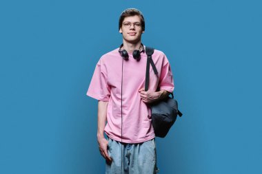 Örgü pembesi tişörtlü, kulaklıklı, mavi renkli stüdyo arkaplanındaki kameraya bakan genç hippi erkek. Gençlik, moda, eğlence, yaşam tarzı konsepti