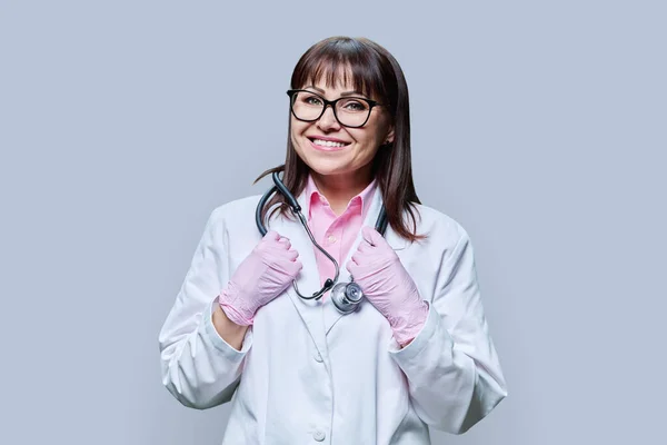 白色实验室外套手套听诊器中中年女医生的画像 在灰色工作室背景下看着相机 医药健康保护 专业医务工作者 医疗服务概念 — 图库照片