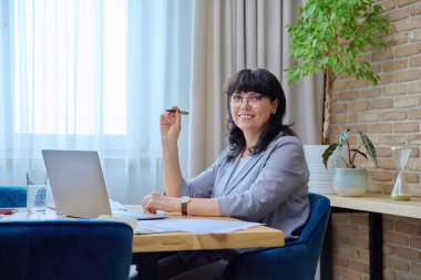 Kendine güvenen, gülümseyen, olgun bir kadın, dizüstü bilgisayarla iş sözleşmesi yapıyor, ofisteki kameralara bakıyor. 50 'lerin kadın lider yöneticisi finans müdürü iş yeri