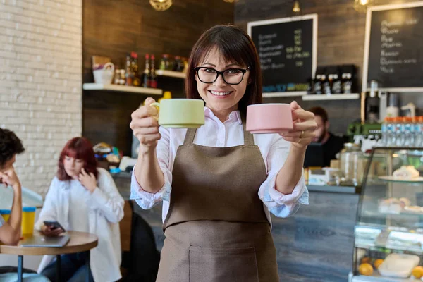 2杯のコーヒーを手にした笑顔の女性コーヒーショップ労働者の姿 カフェホール内のカメラを見てエプロンの女性 小さなビジネス スタッフのコンセプト — ストック写真